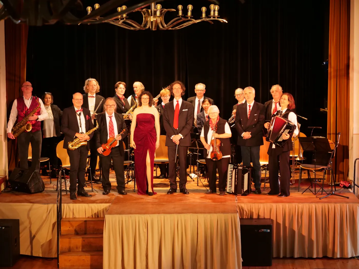Karin Stein im roten Abendkleid mit dem Dirigenten und den Musikerinnen und Musikern des Salonorchesters 1923 auf der Bühne